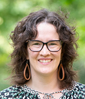 Rosie, a middle-aged white person with tousled shoulder-length hair and glasses, smiles at the camera.
